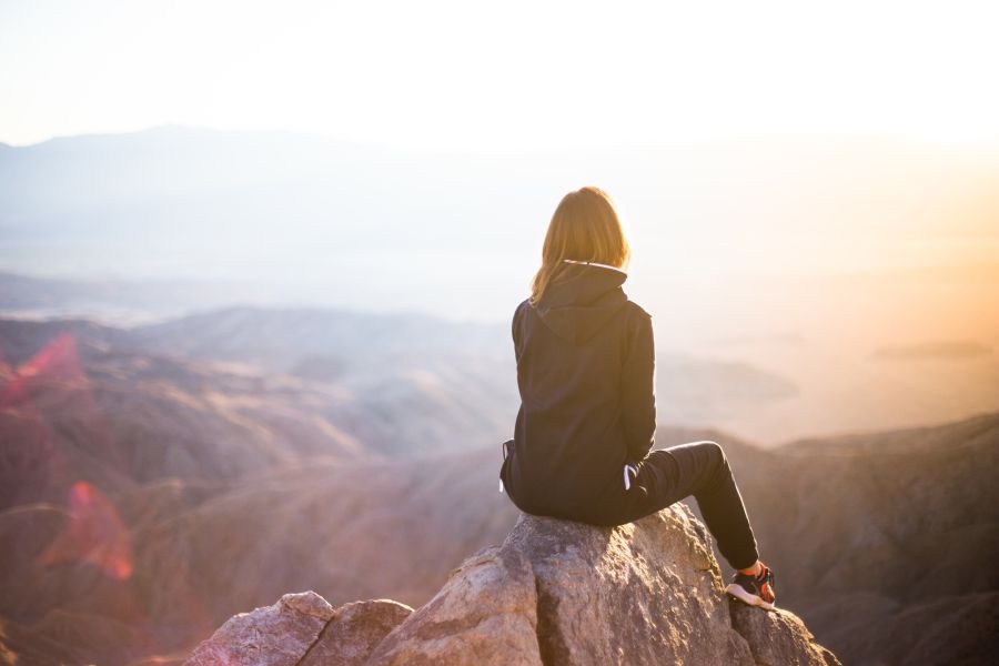Sportlich gekleidete Frau sitzt mit dem Rücken zum Betrachter auf einem Felsen hoch oben über einem canyonartigen Gelände